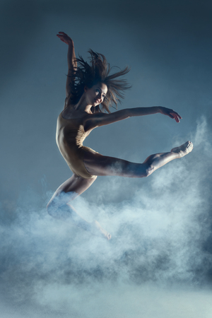 Dancing in cloud concept. Muscle brunette beauty female girl adult woman dancer athlete gymnast in smoke fog wearing dance bodysuit jumping in mid air, performance on isolated grey / black backgroundの写真素材