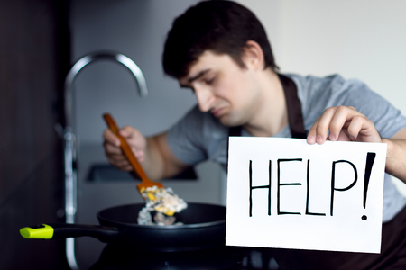 Young loser man male cooking scramble on metal frying pan in modern loft kitchen need help. Trying to make breakfast, lunch or dinner. Alone bachelor in kitchen without woman female conceptの写真素材