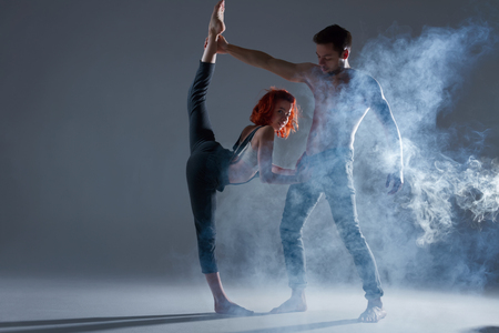 Couple in love dancers making dance element in smoke fume on isolated grey background scene. Redhead woman female dancer athlete gymnast in overalls with muscle male man in jeans and bare naked torsoの写真素材