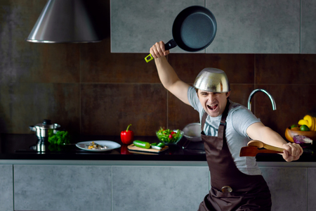 Funny loser man male with metal pan on head frying pan and wooden kitchen utensils in pose of ninja trying to cook failed and yell on grey modern loft kitchen. Unsuccessful bachelor on kitchen conceptの写真素材