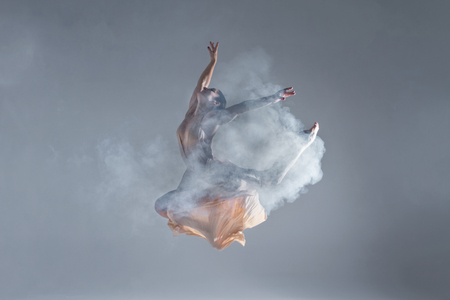 Elegant woman female girl ballerina dancer in beige dress dancing, making performance and dance element in fog dust smoke fume on isolated grey background scene. Dancing in cloud conceptの写真素材