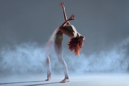 Dancing in cloud concept. Muscle redhead beauty female girl adult woman dancer athlete gymnast in dust fog smoke wearing dance clothing making dance element performance on isolated grey backgroundの写真素材