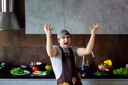 Funny angry loser man male guy with metal sieve on head, frying pan and wooden kitchen utensils trying to cook, failed and yell on grey modern loft kitchen. Unsuccessful bachelor on kitchen concept.の写真素材