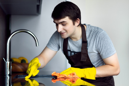 Young man male guy bachelor from clean service with kitchen apron smiling and cleaning kitchen table with cleaning rubber gloves on modern loft black kitchen. Alone man on kitchen conceptの写真素材