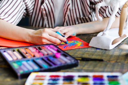 Soiled artists hands draw sketch with pastels on orange sheet of paper. Closeup of creative workplace on wooden antique table with drawing tools, colorful pastels, watercolors.  Art concept.の写真素材