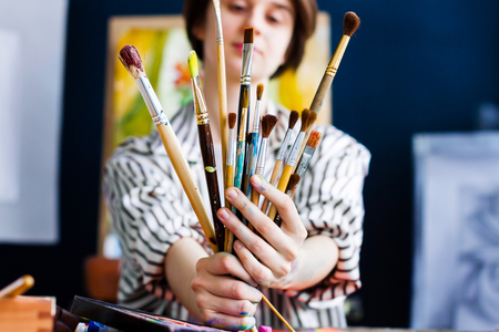 Young beautiful artist teacher student woman girl boy in art workshop studio surrounded by colorful black white painting hold huge bunch stack of art brushes for oil acrylic paints wear striped robeの写真素材