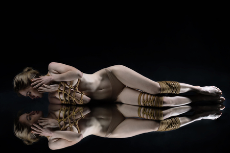Young seductive slim woman lying on mirror floor tied with decorative knots of natural jute rope. Ancient Japanese art of aesthetic bandage and tying shibari kinbaku. Sexual games concept.の写真素材