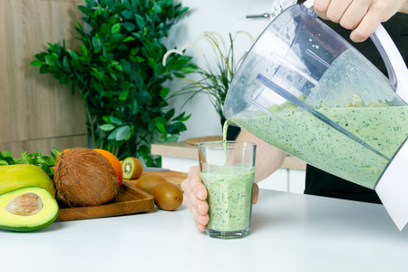 Young man male barista chef with apron doing cook fresh vitamin summer juice smoothie cocktail fruit salad: avocado, mango, kiwi in blender on modern kitchen. Man on kitchen, raw healthy food concept.の写真素材