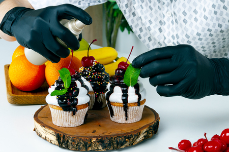 Cook chef men in black gloves prepare decorate splash vegan vegetarian healthy diet sugar lactose free cupcake cake with cherry, chocolate and fresh mint on home wooden kitchen. Food stylist concept.の写真素材