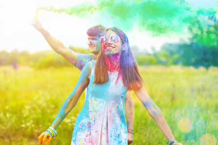 Happy positive smiling fun woman wearing sunglasses all stained with colorful paint celebrating dancing laughs on Holi festival party. On color dust smoke powder cloud background in park field outdoorの写真素材