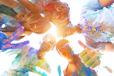 Friends soiled with colorful bright paints, are standing in circle looking down at camera and smiling. Company of young people in sunglasses, having fun with holi paints. Holi party concept.の写真素材