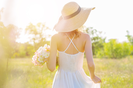 Back view of slim young woman in fashionable hat holding bouquet of pink and white flowers. Romantic girl in white dress is walking and dreaming on spacious field at sunset. Provence style.の写真素材