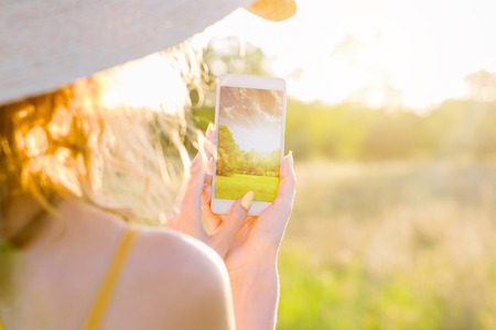 Happy smiling woman female in summer hat and lite yellow summer dress doing picture photo of nature by smartphone, mobile phone in green park outdoor. Summer, spring active outdoor leisure concept.の写真素材