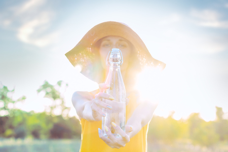 Happy beauty young woman female with pure glass of water wears yellow lite dress and summer hat smiling walking outdoor in green park at sunny warm light day. Active outdoor lifestyle leisure concept.の写真素材