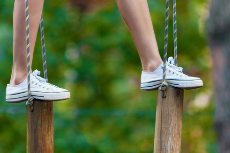 Closeup of female slender legs in white sneakers keep balance on wooden stumps hanging on ropes on trees in park. Rope park with obstacles and ziplines. Extreme rest and summer activities concept.の写真素材