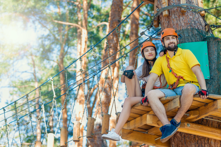 Young woman and man in protective gear are sitting on wooden board on high tree, posing and smiling. Rope adventure park with obstacles and ziplines. Extreme rest and summer activities concept.の写真素材