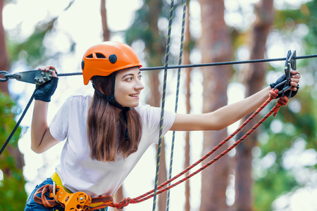 Young beauty female woman adult wears protective helmet having fun in extreme rope park, amusement park. Climbing in rope bridge at green nature forest. Active healthy lifestyle in spring or summer.の写真素材