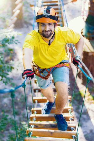 Young male man adult wears protective helmet with action camera having fun in extreme rope park, amusement park. Climbing in rope bridge at green forest. Active healthy lifestyle in spring or summer.の写真素材