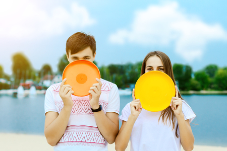 Happy active smiling romantic couple playing disk game Girl boy hiding behind frisbee on sand beech with blue sea sky with clouds Concept of spring summer outdoor amusement activities entertainmentの写真素材