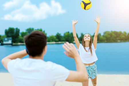 Happy active smiling romantic couple playing volleyball girl hit ball game on sand beech with blue river lake sky with clouds behind Concept of spring summer outdoor amusement activities entertainmentの写真素材