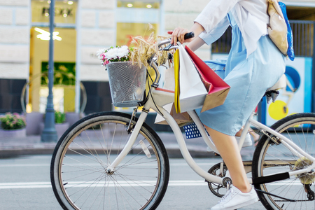 Young woman is cycling around city on white bicycle. Girl in blue dress is going shopping with colorful packages on handlebars of bike. Female retro bicycle with basket with bouquet of flowers.の写真素材