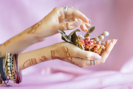Slender elegant female wrists painted with traditional Indian oriental mehndi ornaments by henna. Hands dressed in bracelets and rings hold dry roses flowers. Pink fabric with folds on background.の写真素材