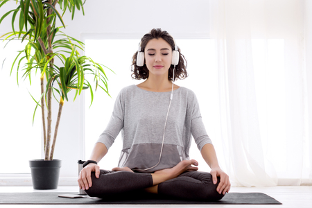 Slender female folded crossed legs in lotus position. Young athletic woman in fitness tracker and headphones is practicing yoga, meditating, relaxing while sitting in padma asana at home.の写真素材