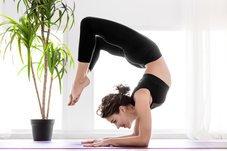 Young slender female is standing at scorpion pose.  Athletic brunette woman in black uniform is...