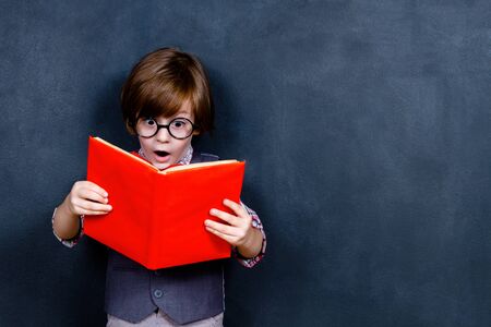 Shocked intelligent smart schoolboy boy with eye glasses reading red book opposite school chalkboard...