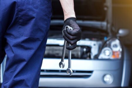 Repairman is repairing car at service station. Back view mechanic hands in gloves are holding steel wrenches. Vehicle with open hood on background.  Modern auto repair shop with equipments and tools.の写真素材