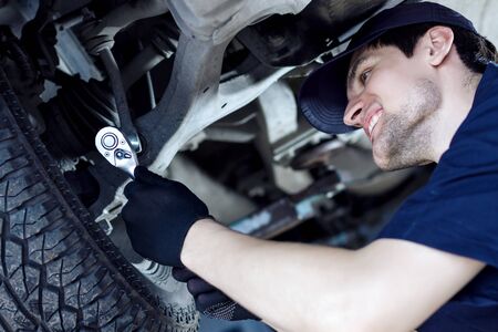 Mechanic in blue jumpsuit is repairing car at service station garage. Smiling repairman is tightening nut by steel wrench at workshop auto repair shop. Vehicle on hydraulic lift is above.の写真素材
