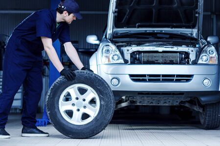 Mechanic in blue jumpsuit is repairing car at modern service station. Repairman is rolling wheel on floor of workshop auto repair shop. Silver vehicle on background. Tire fitting concept.の写真素材