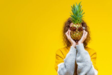 Curly slim girl is holding pineapple dressed in pink glasses in front of her face. Sunny young female woman on yellow background. Bright active lifestyle. Good mood and fun pastime concept.の写真素材
