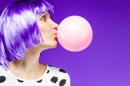 Beautiful girl in violet wig is blowing a huge pink bubble with bubblegum. Stylish fashion woman dressed in blouse with polka dots. Bright young female on purple background. Fun and gladness concept.の写真素材