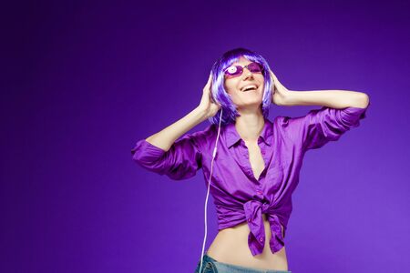 Beautiful smiling girl in wig and headphones is listening music and dancing at party. Stylish fashion woman dressed in violet clothes and glasses. Bright young female on purple background.の写真素材