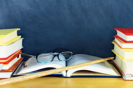 Stacks of colorful books are on either side of desk. Open notebook with glasses and pointer are on center of table in classroom. Chalkboard blackboard on background. Back to school concept.の写真素材