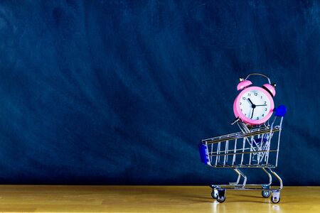 School and study supplies in shopping cart, trolley: pink alarm on wooden desk and chalkboard...