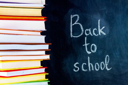 White chalk handwritten inscription of back to school on right side of blackboard. Colorful stack of...