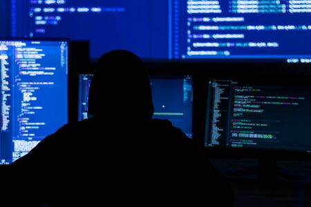 Back view of man in black hood is hacking password. Programmer is writing malware on multiple...