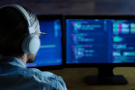 Modern software developer freelancer with white headphones working with program code: C++, Java, Javascript on wide displays at night. Develops new web, desktop, mobile application or framework.の写真素材