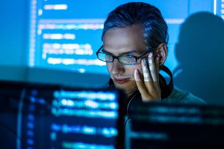 Young tired man freelancer in glasses is working at night because of deadline. Programmer developer...
