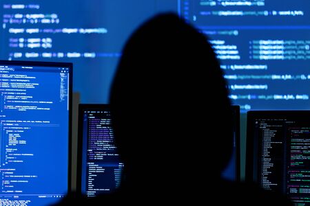 Back view of man in black hood is hacking password. Programmer is writing malware on multiple...
