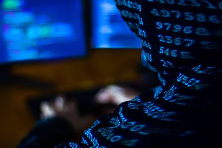 Man in black hood is hacking password. Programmer is writing malware on multiple screens to steal private information. Closeup binary code on his back from projector light. Hacker attack concept.の写真素材