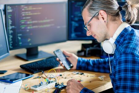 Developer is connecting breadboard to microcontroller. Man is holding smartphone with program code...