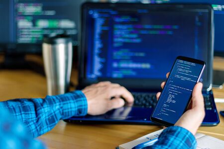 Closeup freelancer hand is holding smartphone with program code. Programmer developer is writing software on multiple computer screens at home office. Devices for working. Geek workplace concept.の写真素材