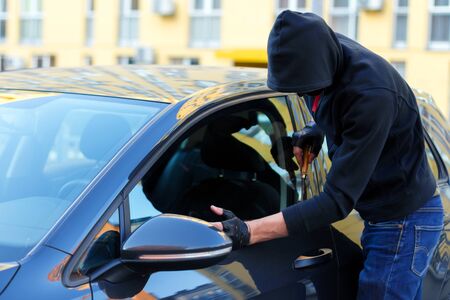 Man thief in black hood and gloves is trying to break into automobile without signaling alarm. Burglar is hacking door lock of private vehicle. Theft in car concept. Bad parking security.の写真素材