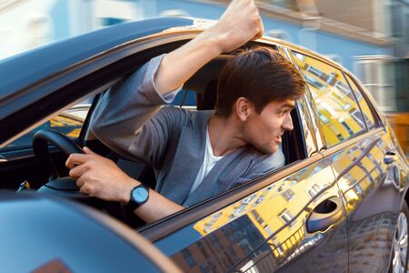 Annoyed young businessman is driving car. Man in gray suit is swearing with other road users....
