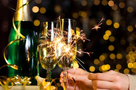 Female hands are holding glass with champagne, burning sparkle. Gold decor, balls, serpentine on table. Decorative garland with yellow light bulbs are shining on background. New year, christmas mood.の写真素材