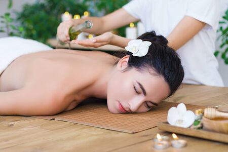 Young pleased woman is getting thai massage, therapy. Female master is pouring massaging oil in palms. Brunette girl is lying on couch in light spa ayurveda salon. Relax and health care concept.の写真素材