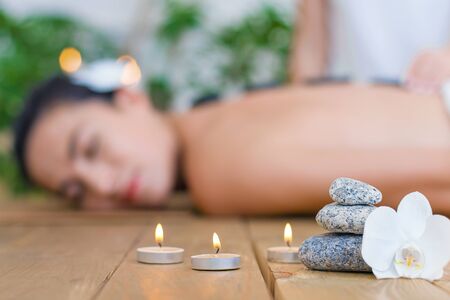 Closeup burning candles, flower, special stones. Young woman is getting hot thai massage, therapy. Brunette girl is lying on couch in light spa ayurveda salon. Relax and health care concept.の写真素材